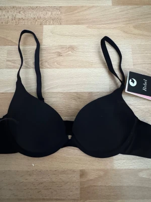 Ny. Svart push-up bh från Rebel - Stilren svart push-up bh från Rebel med släta kupor och justerbara axelband. Bh:n har bygel för extra stöd och en enkel design utan spets eller dekorationer. Materialet känns mjukt och bekvämt mot huden.