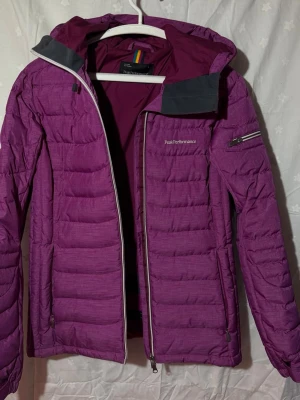 Lila dunjacka från Peak Performance - Snygg lila dunjacka från Peak Performance med huva och dragkedja. Jackan har quiltad design, ficka på ärmen och diskret logga på bröstet. Perfekt för kalla vinterdagar och riktigt varm tack vare dunfyllningen. Snölås och ficka för skidkort.