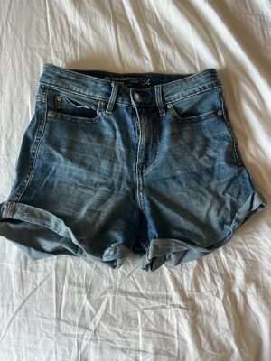 Blå denimshorts med uppvik - Klassiska blå jeansshorts med hög midja och uppvikta benslut. Femficksmodell med knapp och dragkedja framtill. Perfekta för varma dagar och enkla att matcha med allt.