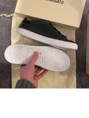 Svarta sneakers från Axel Arigato - Stilrena svarta sneakers från Axel Arigato med lågt skaft och vit sula. Skorna har en minimalistisk design med svarta skosnören och är tillverkade i slätt läder. Perfekta för dig som gillar en clean och modern look.