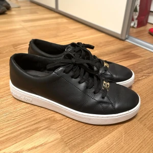 Svarta sneakers från Michael Kors 39 - Snygga svarta sneakers i skinn från Michael Kors med vita sulor och guldfärgade detaljer. Skorna har snörning och en stilren design som passar perfekt till vardags. Michael Kors-loggan är präglad på sidan av sulan. Storlek 39. Skorna är använda men i bra skick som ni kan se. Köpte för 1595:-