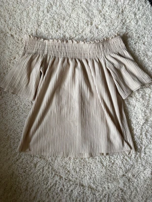 Beige offshoulder topp Gina Tricot - Säljer en beige offshoulder topp från Gina Tricot med smock upptill och korta, vida ärmar. Toppen har en ribbad struktur och är perfekt för varma dagar. 