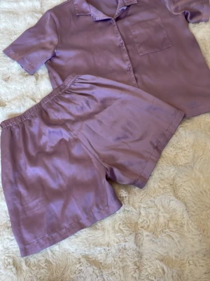 Lila satinpyjamas från Lindex - Snygg lila pyjamas i satin från Lindex  . Setet består av en kortärmad skjorta med krage, knappstängning och bröstficka samt matchande shorts med resår i midjan. Tillverkad av återvunnen polyester för en mjuk och glansig känsla. Storlek 146/152. Pris 30kr