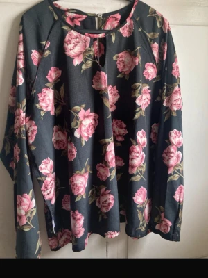 Blommig långärmad blus - Blus med stora rosa blommor och gröna blad. Blusen har rund halsringning och långa ärmar. Materialet ser ut att vara mjukt och lätt, perfekt för en trendig och avslappnad look.