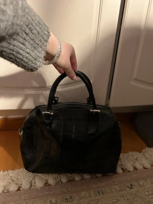 Svart handväska i crocopräglat skinn - Snygg svart handväska i crocopräglat konstläder med två handtag och silverfärgade detaljer. Väskan har dragkedja upptill och rymligt huvudfack. Väskan är passlig storläk till exempel till skolan och den har två fickor! Jag har köpt den använd så vet inte märket.