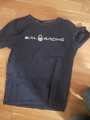 Mörkblå Sail Racing t-shirt - Snygg mörkblå t-shirt från Sail Racing med vit logga tryckt på bröstet. Klassisk rund halsringning och korta ärmar. Tillverkad i mjuk bomull som känns skön mot huden. Perfekt för dig som gillar stilrena och sportiga plagg.