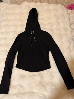 Svart ribbad hoodie från Stronger - Snygg svart hoodie från Stronger med ribbad struktur och långa ärmar. Huvan har dragsnören med loggan 'STRONGER' tryckt i vitt. Perfekt för en chill och sportig look. Materialet känns mjukt och passar bra till träning, och modellen är något croppad.