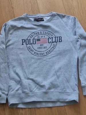 Grå sweatshirt från Polo Club - Snygg ljusgrå sweatshirt från Polo Club med rund halsringning och tryck framtill med amerikansk flagga och text. Tröjan har långa ärmar och ribbade muddar vid ärmslut och nederkant. Perfekt för en avslappnad och sportig stil. Syl M
