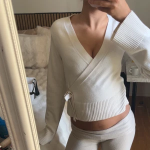 Vit omlottkofta från Gina Tricot - Snygg vit omlottkofta från Gina Tricot med långa ärmar och ribbade muddar. Koftan har knytband i midjan och en djup v-ringning som ger en trendig look. Perfekt att styla med både jeans och kjol för en fräsch outfit.