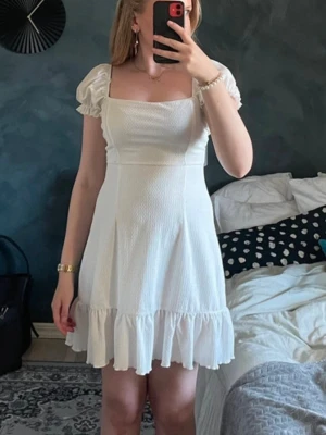 Vit kort klänning med snörning - Söt vit kortklänning med puffärmar, fyrkantig urringning och volangkant nertill. Klänningen har öppen rygg med snygg snörningsdetalj. Perfekt för avslutningar/student och konfirmation. 