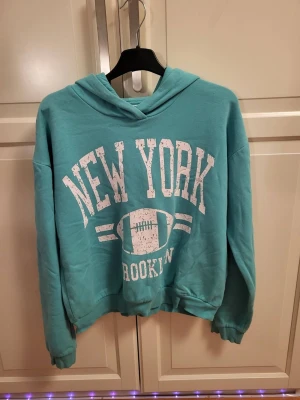 Turkos hoodie med New York-tryck - Snygg turkos hoodie med vit text 'NEW YORK' och 'BROOKLYN' samt en amerikansk fotboll på bröstet. Hoodien har huva och ribbade muddar vid ärmslut och nederkant. Perfekt för en avslappnad streetwear-look.
