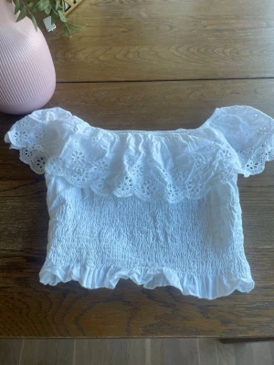 Vit offshoulder blus med volang - Superfin vit offshoulder blus med broderade volanger och smockad kropp. Blusen har romantiska detaljer med spets och volangkant nertill. Perfekt för en somrig look och passar till både jeans och kjol. Är från samma sätt som den vita kjolen.