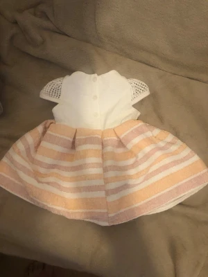 Söt kort klänning med spetsärmar för en tjej  - Supersöt kort klänning med vit överdel, virkade spetsärmar och kjol i ljusrosa och vita ränder. Kjolen är lätt utställd och har en mjuk struktur. Perfekt för sommarens alla tillfällen! Den är st 86 till ett år.