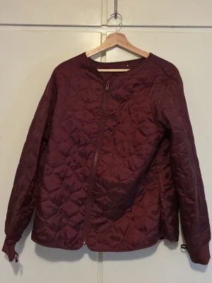 Vinröd quiltad pufferjacka - Stilren vinröd pufferjacka med quiltat mönster och dragkedja framtill. Jackan har rund halsringning och långa ärmar. Perfekt för kalla dagar och ger en trendig look. Materialet är glansigt och ger en modern känsla.