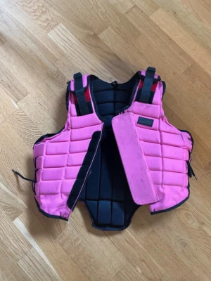 Rosa ridväst från Hööks - Säljer denna rosa ridväst grån Hööks i stl 12. Den är i bra skick, har lite fläckar (som jag inte vet om de går bort eller inte), men inget som förstör användningen. Den är 47 cm uppifrån och ner, se sista bilden! Hör av er om frågor och kontakta mig innan köp💗(Säljer även på Vinted ifall man skulle vilja köpa där)