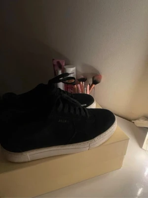 Svarta sneakers från Axel Arigato - Stilrena svarta sneakers från Axel Arigato med låg profil och vit sula. Skorna har en klassisk siluett och är tillverkade i mocka, vilket ger en lyxig känsla. Perfekta för dig som gillar minimalistisk design och vill ha något som funkar till det mesta.