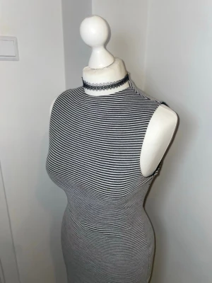 Randig ärmlös klänning med hög hals - Snygg kroppsnära klänning med svartvita ränder och hög hals. Klänningen är ärmlös och har en dragkedja i ryggen. Perfekt för dig som gillar en stilren och modern look. Materialet är mjukt och stretchigt, vilket gör den bekväm att bära.