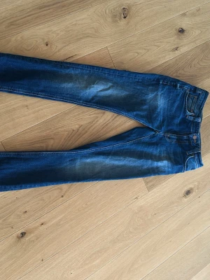 Mörkblå nudie slim fit jeans - Säljer ett par mörkblå slim fit jeans med snygga slitningar och tvättade detaljer på låren. Klassisk femficksmodell med knapp och dragkedja. Perfekta till en avslappnad stil och passar till det mesta. Materialet är mjukt och stretchigt för extra komfort.