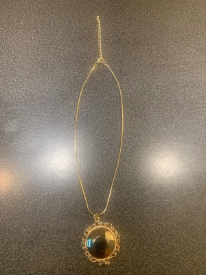 Guldhalsband med stor grå sten - Snyggt halsband med tunn guldfärgad kedja och ett stort, facettslipat svart hänge. Hänget är runt och omges av små guldfärgade detaljer och grå strasstenar som ger en vintagekänsla. Perfekt för dig som vill ha ett statement-smycke med unik design.