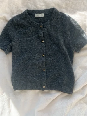 Mörkgrå kortärmad kofta H&M - Stl 158/164.Kortärmad kofta i mjuk mörkgrå ullmix från H&M. Rund halsringning, ribbade muddar och knäppning med små pärlemorknappar framtill. Perfekt att styla över en topp eller klänning för en chill look.