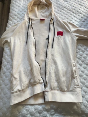 Vit hoodie med dragkedja från Hugo - Säljer en stilren vit hoodie från Hugo med dragkedja framtill och svart snörning i huvan. På bröstet sitter en röd patch med Hugo-loggan. Hoodien har ribbade muddar och är tillverkad i mjukt bomullsmaterial.