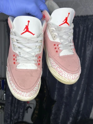 Rosa och vita Air Jordan 3 sneakers - Säljer ett par Air Jordan 3 sneakers i rosa och vitt med röda detaljer och klassiskt Jumpman-logga på plösen och hälen. Skorna har vita snören, rosa mockaliknande material och mönstrad sula. Perfekta för dig som vill sticka ut med en unik färgkombo.