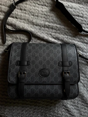 Väska från Gucci, Gucci Messenger bag suprime canvas  - Snygg svart axelväska från Gucci med klassiskt GG-mönster och justerbar axelrem. Väskan har två spännen framtill och präglad logga på locket. Tillverkad i slitstarkt konstläder med detaljer i svart läder. Perfekt för dig som gillar stilrena accessoarer.