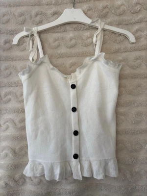 Vit ribbad linnetopp med volang - Supersöt vit linnetopp med knytbara axelband, svarta knappar framtill och volangkant både upptill och nedtill. Toppen är ribbad och har en feminin, somrig look som passar perfekt till jeans eller kjol. Den har inget märke eller storlek så om ni vill veta måtten kan ni skriva privat!❤️