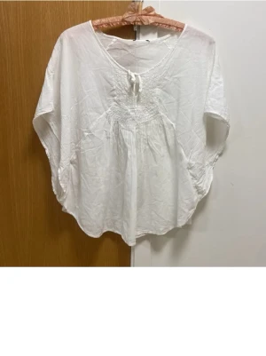 Vit boho blus med knyt - Luftig vit blus i boho-stil med v-ringning och knyt framtill. Blusen har vida, korta ärmar och ett smockat parti över bröstet som ger en avslappnad och flowy känsla. Perfekt till sommaren och enkel att styla med shorts eller jeans.