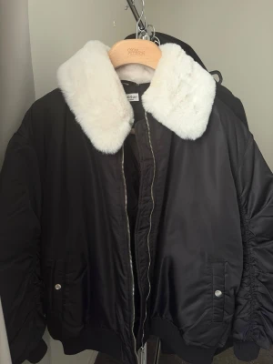 Svart bomberjacka med fuskpälskrage - Snygg svart bomberjacka från Doom Sayers med stor vit fuskpälskrage. Jackan har dragkedja framtill, två fickor med tryckknappar och rynkade detaljer längs ärmarna. Tillverkad i glansigt syntetmaterial och har ribbade muddar vid ärmslut och nederkant.