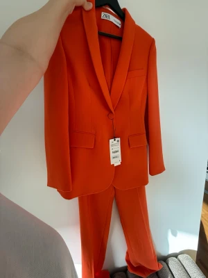 Orange kostym från Zara - Snygg orange kostym från Zara med klassisk kavaj och matchande byxor. Kavajen har en knapp, två fickor med lock och en bröstficka. Kostymen har en modern passform och är tillverkad i ett slätt syntetmaterial. Kavajen kan säljas separat eller med byxorna. Nypris för kavajen 859:- och byxorna 399:- 