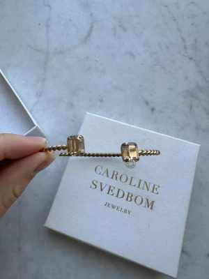 Guldarmband Caroline Svedbom - Snyggt armband i guldton från Caroline Svedbom med tvinnad design och två stora, rektangulära kristaller i båda ändar. Armbandet är öppet och har en exklusiv känsla med gnistrande detaljer som verkligen sticker ut.