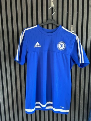 Chelsea FC Adidas blå träningströja (köpt i London) - Snygg blå Chelsea FC träningströja från Adidas med vita detaljer och klubbmärke på bröstet. Tröjan har klassiska Adidas-ränder på axlarna, rund hals och korta ärmar. Perfekt för fotbollsträning eller att bära till vardags.