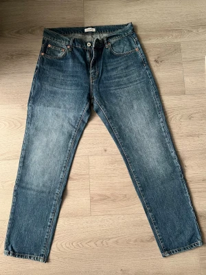 Blå raka jeans från Woodbird - Säljer ett par klassiska blå jeans från Woodbird med rak passform och fem fickor. Jeansen har snygga slitningar och kontrastsömmar, tillverkade i slitstark denim. Perfekta för en avslappnad och trendig look.