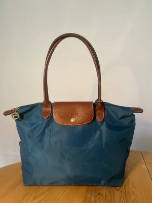 Klassisk Longchamp Le Pliage handväska - Stilren blå/turkos handväska från Longchamp Le Pliage med bruna läderdetaljer och handtag. Väskan har guldfärgade metalldetaljer och dragkedja med Longchamp-logga. Tillverkad i slitstark nylon och äkta läder. Perfekt för dig som gillar klassisk och enkel design. Väskan är i fint skicka och fungerar perfekt. Dragkedja och knapp fungerar som de ska. Ca mått Bredd: ca 28-30, Höjd: ca 26-28 cm, Djup: ca 15 cm. Handtag ca 21 cm.