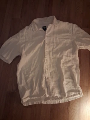 Vit linneskjorta från FSBN - Klassisk vit skjorta från F&F i svalt linne. Skjortan har korta ärmar, bröstficka och knappar framtill. Perfekt för varma dagar och enkel att styla till olika outfits.