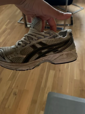 Asics vita sneakers med mesh och svarta detaljer - Säljer ett par vita Asics sneakers med svart logga och detaljer. Skorna har en sportig design med mesh-ovandel för bra ventilation och syntetiska förstärkningar. Klassisk snörning och rejäl sula för komfort. Perfekta för dig som gillar retrostil och streetwear.