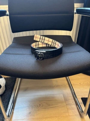 Burberry bälte med klassiskt rutmönster - Säljer ett Burberry bälte med den ikoniska rutiga insidan och svart utsida i skinn. Spännet är i metall med Burberrys TB-logga. Perfekt accessoar för att lyfta din outfit med en lyxig touch.