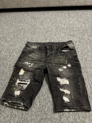 Svarta jeansshorts från Dsquared2 - Säljer ett par svarta jeansshorts från Dsquared2 med slitningar och coola färgstänk i gult och vitt. Shortsen har fem fickor, knappgylf och en längre modell som går till knäna. Perfekta för dig som gillar streetwear och edgy stil.