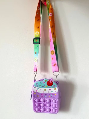 Lila cupcake handväska med pop it - Söt handväska i form av en cupcake med pop it-detaljer i lila silikon. Väskan har färgglad axelrem med justerbar längd och mönster av donuts och godis. Dragkedja upptill och dekorativt körsbär på toppen. Perfekt för dig som gillar lekfulla accessoarer.ny ej använd 