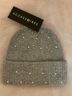 Grå pärlprydd mössa från Accessoires - Snygg grå ribbstickad mössa från Accessoires, dekorerad med små vita pärlor och glittriga stenar för en extra stylish look. Perfekt för att lyfta vilken outfit som helst under kalla dagar. Mjuk och skön kvalitet.