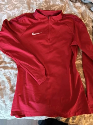 Röd långärmad träningströja Nike - Röd långärmad träningströja från Nike med halv dragkedja framtill och vit logga på bröstet. Tröjan har en sportig passform och är tillverkad i ett mjukt, stretchigt material som passar perfekt för träning eller uppvärmning.