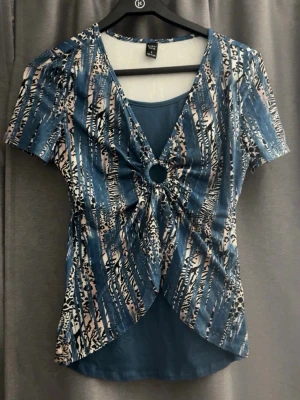 Blå mönstrad omlottopp med ringdetalj - Trendig blå omlottopp  med korta ärmar och coolt abstrakt mönster i blått, vitt och svart. Toppen har en snygg ringdetalj framtill och v-ringad hals. Perfekt för dig som vill sticka ut lite extra.