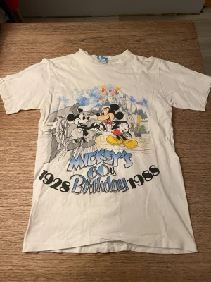 Vit Mickey Mouse 60th Birthday t-shirt - Vintage t-shirt i vitt med stort färgglatt tryck av Mickey Mouse och texten 'Mickey's 60th Birthday 1928-1988' på framsidan. Klassisk passform med korta ärmar och rund hals. Perfekt för dig som älskar retro och Disney vibes.