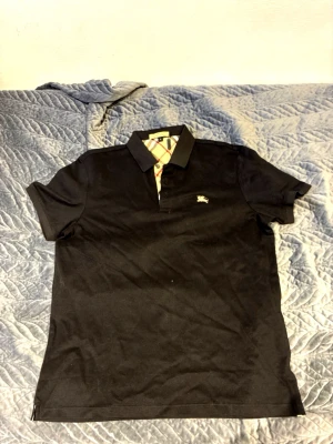 Svart Burberry piké med rutig krage - Snygg svart pikétröja från Burberry med klassisk rutig detalj sop syns vid kragen. Ikonisk Burberry-logga på bröstet och mjukt bomullsmaterial. Perfekt för dig som vill ha en stilren och tidlös look. Är XL men är mer som en M