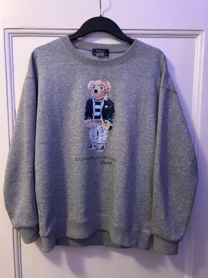 Grå sweatshirt Polo Bear Ralph Lauren - Säljer en grå sweatshirt från Polo Ralph Lauren med den ikoniska Polo Bear-trycket på bröstet. Tröjan har rund halsringning, ribbade muddar och är gjord i mjukt bomullsmaterial. Perfekt för en avslappnad och stilren look.