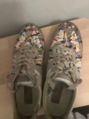 Maison Margiela sneakers med färgstänk - Unika sneakers från Maison Margiela i vitt och beige läder och mocka, dekorerade med färgstänk i rött, blått, gult och svart. Klassisk låg modell med gummisula och beige snörning. Perfekta för dig som vill sticka ut med din stil. Defekt på sista bild!