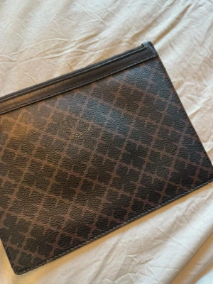 Brun mönstrad kuvertväska i skinn - Snygg brun kuvertväska i skinn med mörkbrunt grafiskt mönster och dragkedja upptill. Väskan har en avtagbar handledsrem och detaljer i guld. Perfekt för att bära det viktigaste och ger en stilren touch till din outfit.