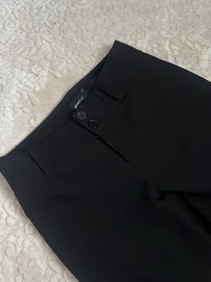 Svarta kostymbyxor från Ginatricot - Snygga svarta kostymbyxor från Ginatricot med raka ben och hög midja. Klassisk design med bälteshällor, knapp och dragkedja framtill. Perfekta till en stilren look och enkla att matcha med olika toppar. Köpt för 500kr