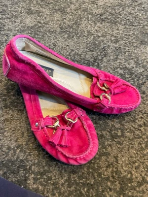 Rosa mockaloafers från Novita - Snygga rosa loafers i mocka från Novita med dekorativa tofsar och guldiga spänne framtill. Skorna har en klassisk form och detaljerade sömmar runt tån. Perfekta för dig som vill sticka ut med färg och stil. De har blivit lite platta för saker har tryckt ner de men inget som syns eller på märks när man har på de!